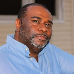 A black man in a blue shirt leaning against a wall.